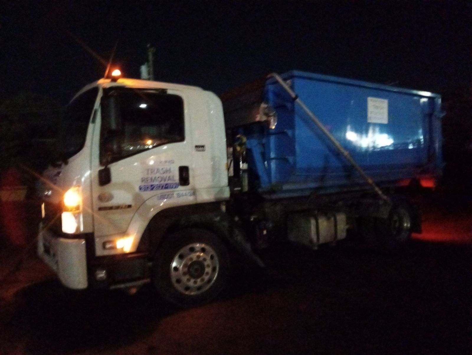 A white roll-off dump truck with a large blue container attached, parked at night with its headlights on.