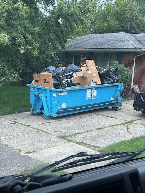 A bright blue dumpster overflowing with trash and cardboard boxes sits in the driveway of a brick house.