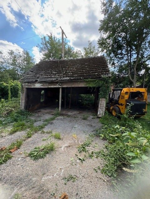 An old, weathered garage with a dark, mossy roof sits on a gravel lot next to a yellow skid-steer loader.