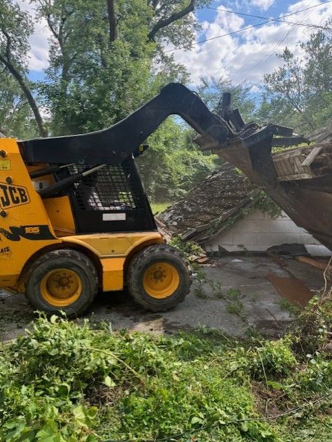 A yellow JCB skid steer loader uses its front attachment to move debris and fallen tree branches outdoors.