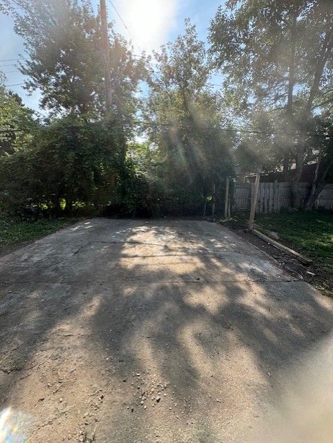 A view of a weathered concrete driveway leading toward dense trees and a wooden fence under a bright, sunny sky.