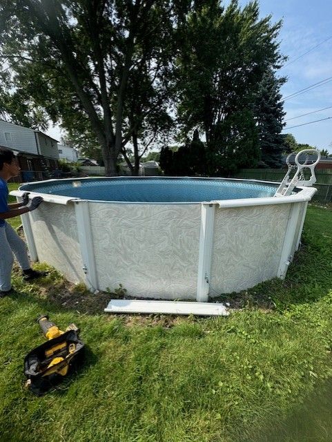 A person in a blue shirt installing white top rails on a round, above-ground swimming pool in a grassy backyard.