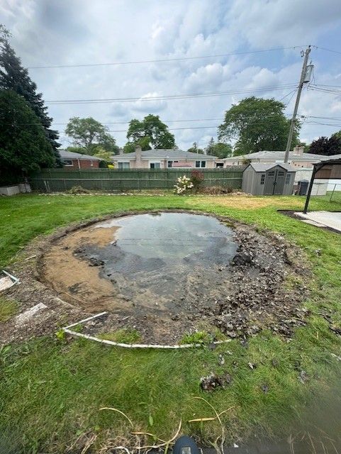 A flooded circular patch of dirt and mud in a backyard, bordered by grass and a fence under a cloudy sky.