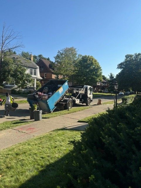 A truck with a blue dumpster is parked on a suburban street, with a person nearby moving trash containers.