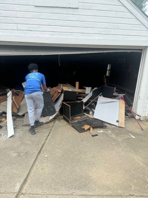 A person in a blue shirt and light pants moves debris out of a garage filled with construction materials and waste.