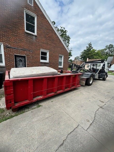 A large red dumpster holding a mattress sits on a concrete driveway next to a brick house and a white roll-off truck.