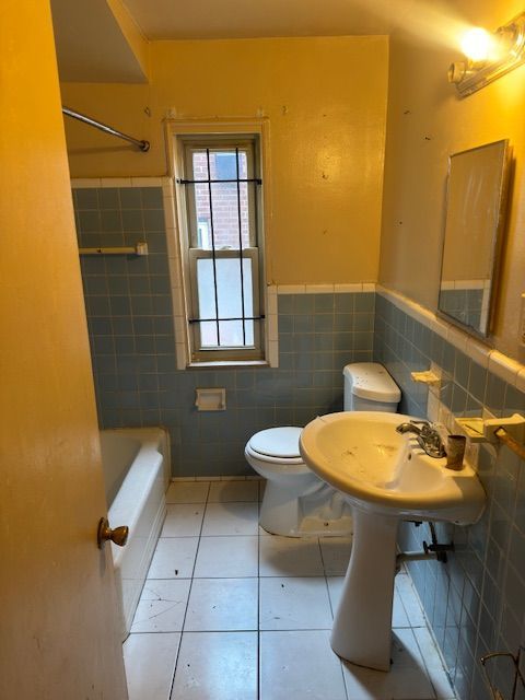 A small, dated bathroom with blue-tiled walls, a pedestal sink, white toilet, and a bathtub under a window.