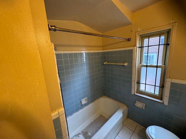 A bathroom with blue tile walls, a white bathtub, a shower curtain rod, a window with a metal grate, and yellow walls.