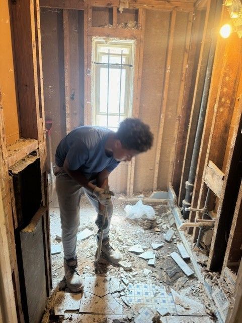 A person in work clothes uses a small hand tool to clear debris from the floor of a room undergoing demolition.