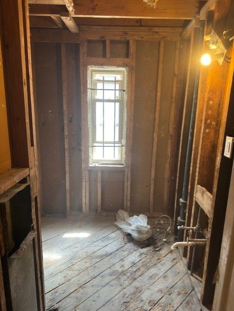 A bare, gutted bathroom undergoing renovation with exposed wooden wall studs, wood plank floors, and a small barred window.