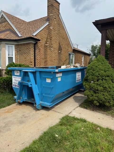 A large, bright blue dumpster sits on a concrete driveway in front of a brick house with a green pine tree nearby.