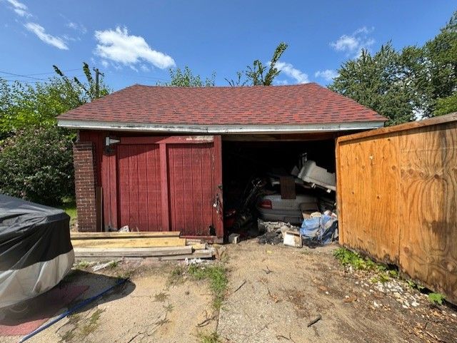A red garage with an open door filled with stored items, next to a plywood fence under a blue sky.
