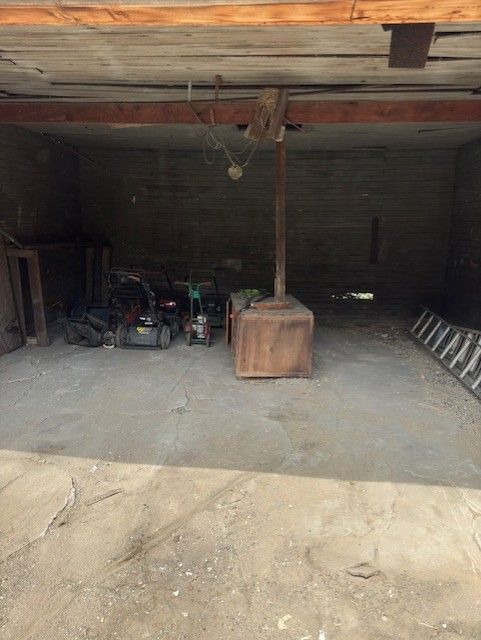 A dimly lit, unfinished garage with a concrete floor, a wood-burning stove, lawn equipment, and a leaning ladder.