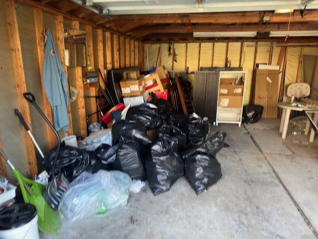 A cluttered garage interior with numerous black trash bags piled on the floor, boxes, and a small table in the corner.