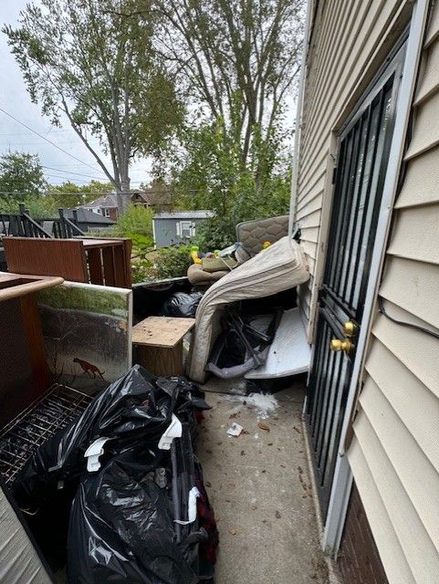 A cluttered outdoor porch piled with trash bags, a mattress, and various discarded furniture against a house wall.