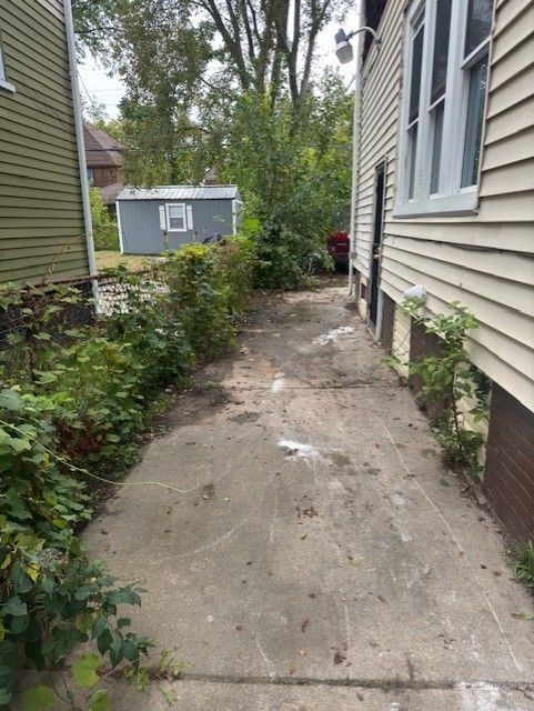 A concrete pathway runs between a green house and a beige house, leading toward a small shed and trees in the background.
