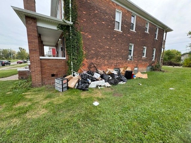 A brick apartment building with a pile of trash bags and cardboard boxes dumped against the side of the exterior wall.