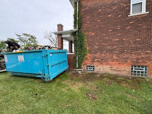 A bright blue dumpster sits on the grass beside a brick house covered in green vines near a porch.