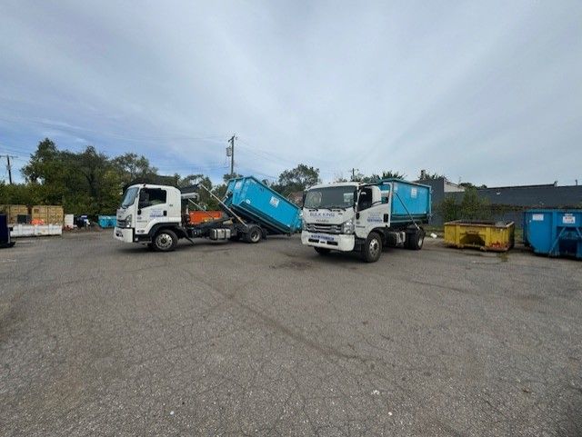Two white trucks with blue dumpsters parked in an outdoor gravel lot under a cloudy sky.