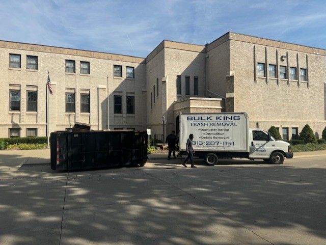 Two people stand near a Bulk King trash removal truck parked in front of a brick building next to a large, tipped dumpster.