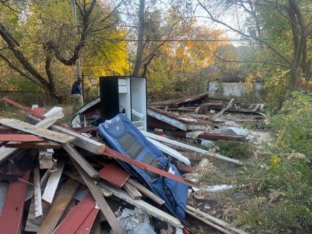 Debris and a blue chair sit in front of a demolished building in a wooded area with a person standing in the background.
