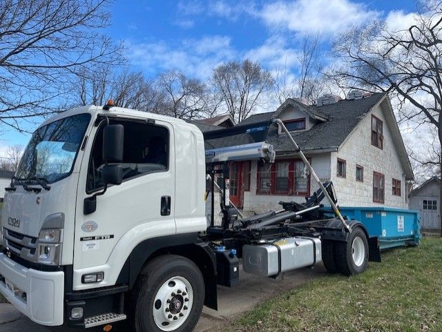 A white Isuzu roll-off truck parked in front of a white house with a blue dumpster on its bed.