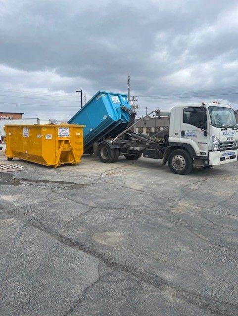 A white roll-off truck loading a large blue dumpster next to an empty yellow dumpster on an asphalt lot under a cloudy sky.