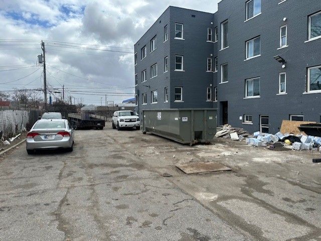 An alleyway beside a dark gray apartment building with parked cars, a large dumpster, and debris on the ground.