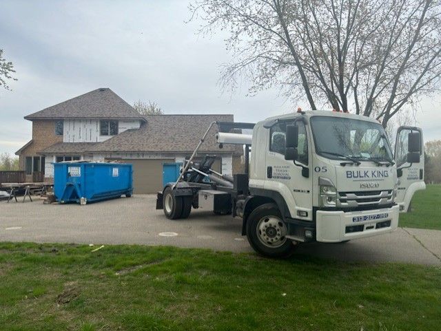 A white Bulk King roll-off truck is parked in a residential driveway next to a blue dumpster at a house under construction.
