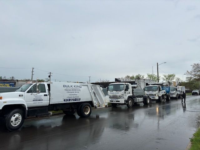 A line of white dump trucks parked on a wet, paved road under an overcast sky.