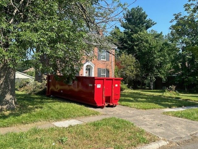 A large, red dumpster sits in the front yard of a red brick house under a leafy tree on a sunny day.