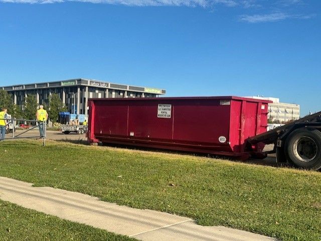 A large red roll-off dumpster sits on a grassy area next to a sidewalk with a parked truck and two people standing nearby.