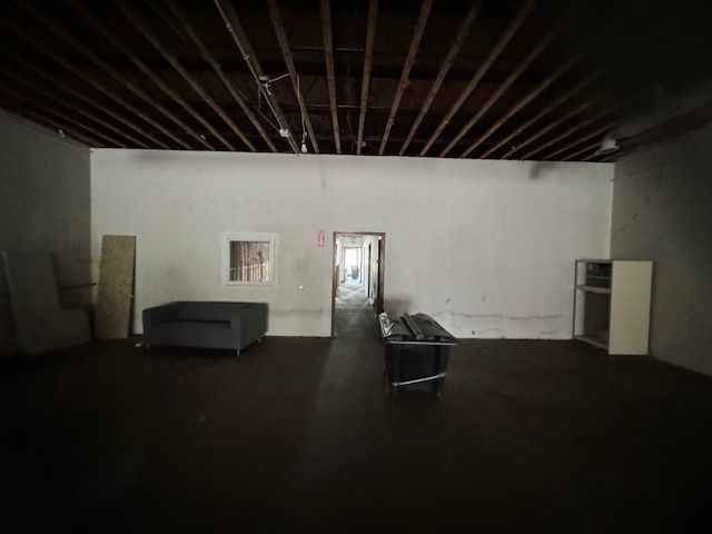An empty, dimly lit room with a wooden rafter ceiling, two dark sofas, a small window, and a doorway to another room.