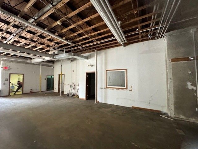 An unfinished, industrial-style room with exposed wooden rafters, white walls, concrete floors, and multiple doorways.
