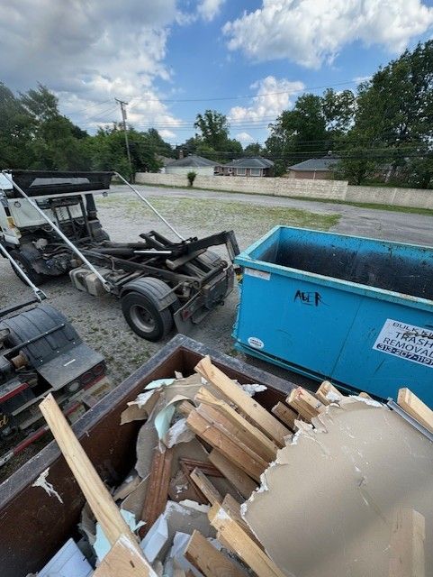 A flatbed truck parked near a bright blue dumpster in a gravel lot with construction debris in the foreground.