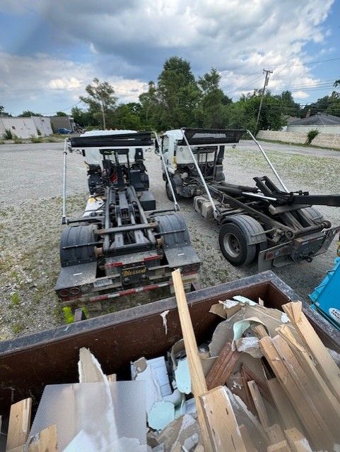 Two roll-off dumpster trucks parked on a gravel lot, viewed from above a debris-filled container.