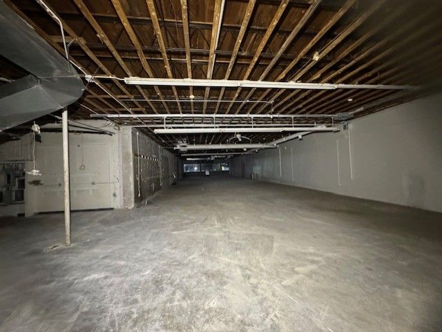 A wide, empty commercial space with concrete floors, white walls, and an exposed wood beam ceiling with hanging lights.