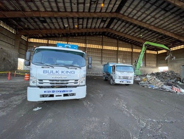 Two white dump trucks and a green excavator inside a large, open-air warehouse facility with industrial debris.
