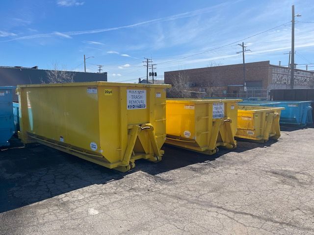 Several yellow and blue dumpsters are lined up in an outdoor gravel lot on a sunny day.