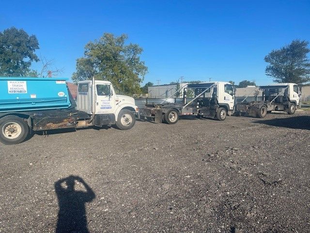 Three white roll-off trucks parked in a gravel lot under a clear blue sky, one carrying a blue dumpster.