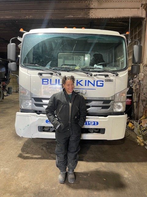 A smiling individual in dark work coveralls stands in a garage in front of a white truck labeled