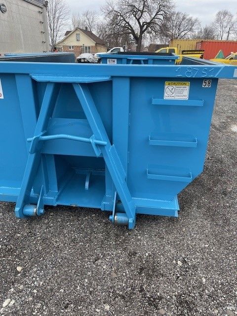A blue metal roll-off dumpster sits on a gravel lot with a residential house and other vehicles in the background.