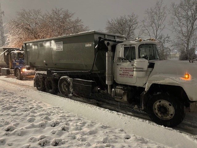 A white roll-off dump truck parked on a snowy roadside during a snowfall.