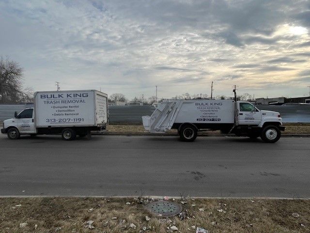 Two white Hulk Kind trash removal trucks parked on a road against a cloudy sky.