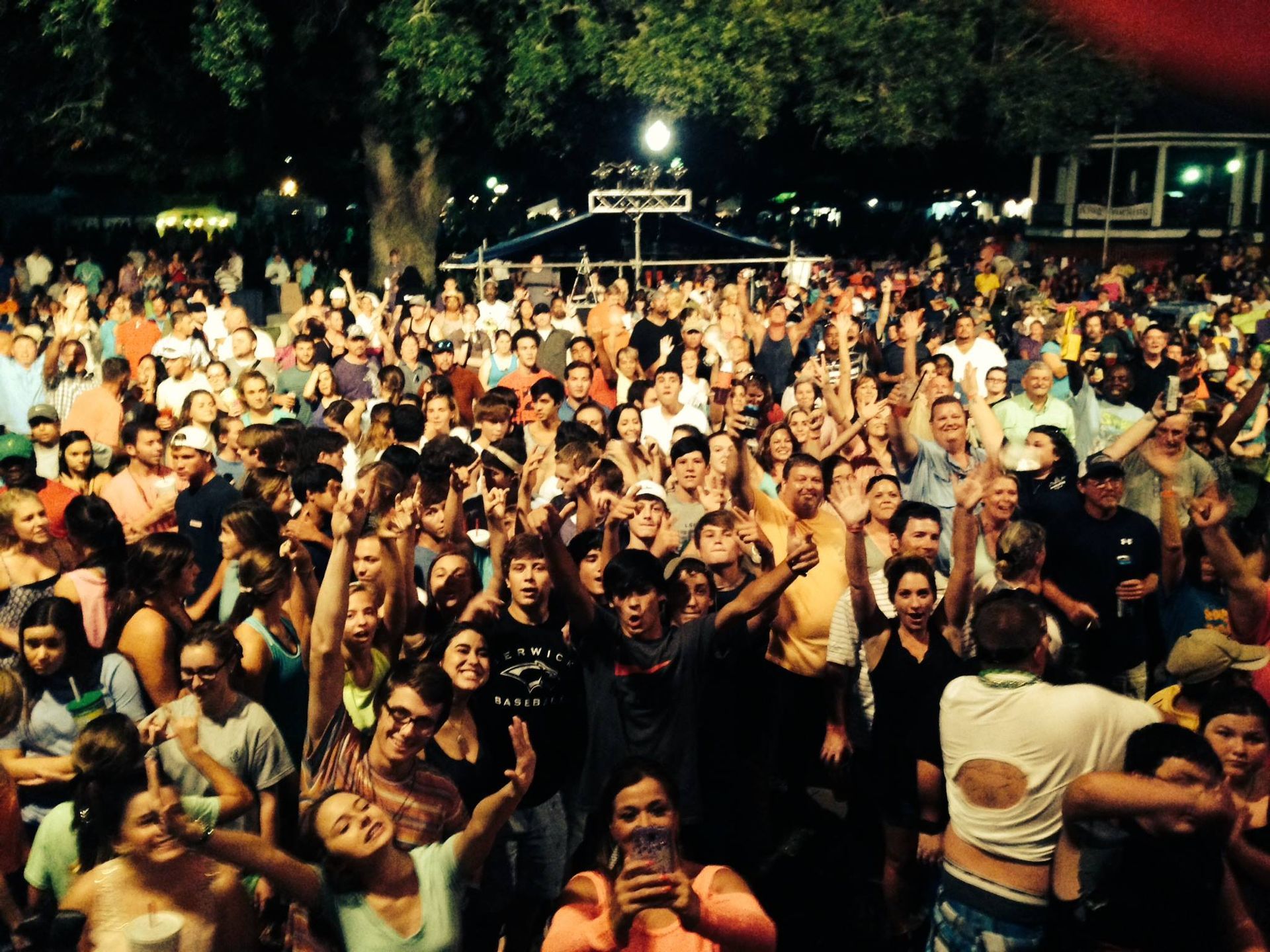 Crowded outdoor concert audience cheering and raising hands at night under warm stage lights