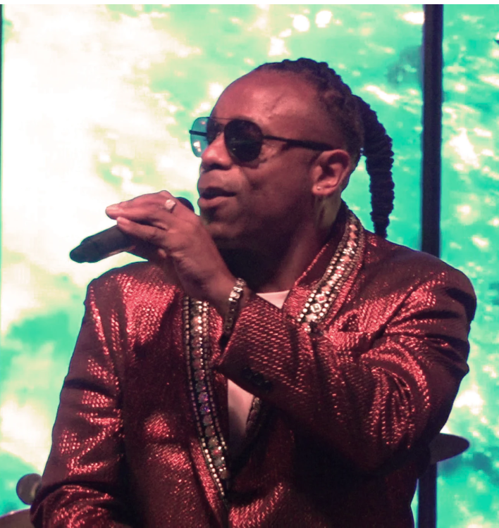 Singer in red sequined jacket and sunglasses holding a microphone on stage