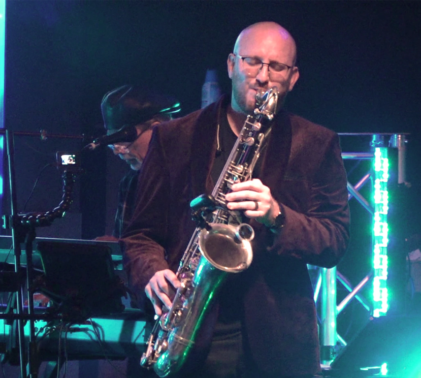 Saxophonist performing onstage under blue and green lights, with a pianist in the background