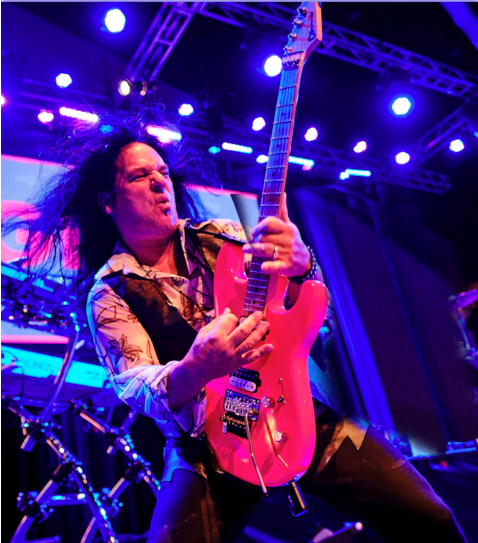 Guitarist performing on stage with a bright pink electric guitar under purple lights