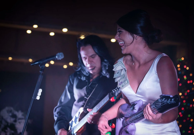 Two musicians performing on stage, smiling under string lights, one in a white dress holding a guitar.