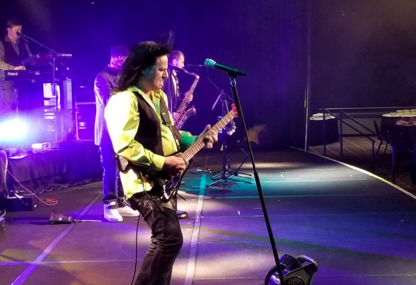 Guitarist performing on a purple-lit stage, singing into a microphone while playing electric guitar